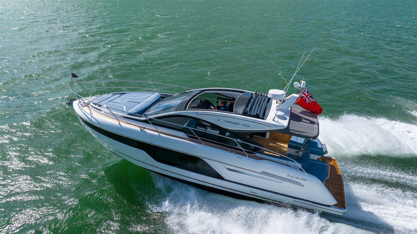 A sleek Fairline Targa 40 yacht glides through turquoise waters, leaving a white wake; a British flag flies from the stern.